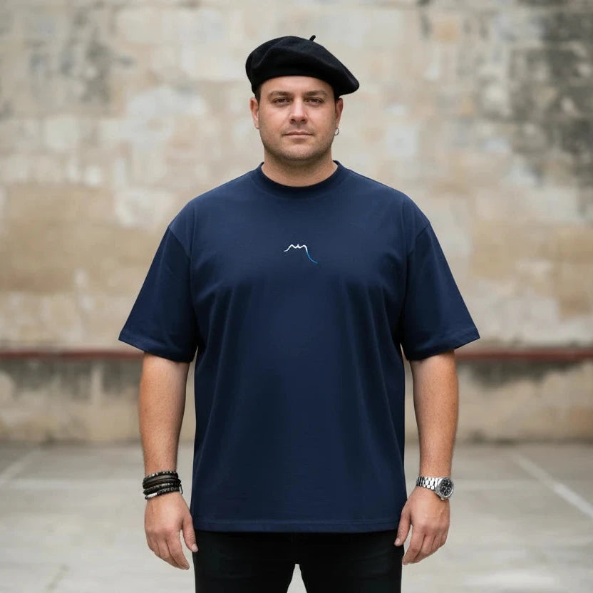 homme portant le t-shirt grande taille en coton bio brodé le pottolo coloris bleu marine devant un fronton au pays basque